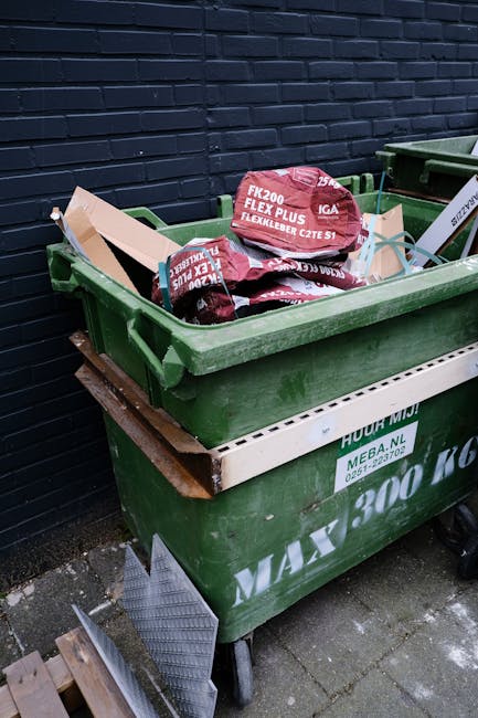 A black plastic wheelie bin with the label 'ST. JOHN'S', positioned on a sidewalk at night, with its lid open and filled with various discarded items including cardboard boxes, paper, and plastic wrap