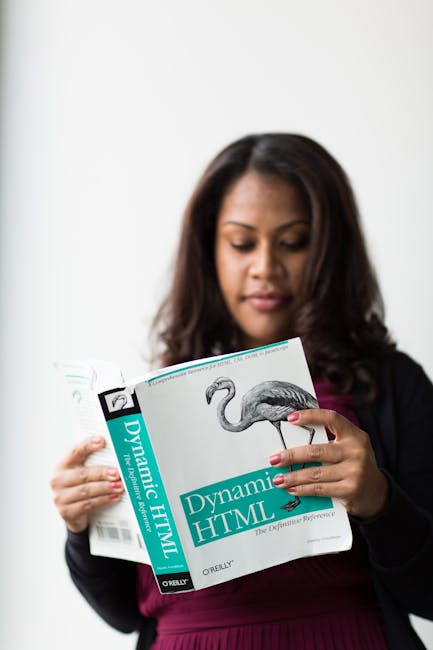 A woman with dark, shoulder-length hair and medium skin tone, dressed in a dark blazer over a maroon top, is holding and reading a book titled 'Dynamic HTML' by ORIELY. She is standing indoors against