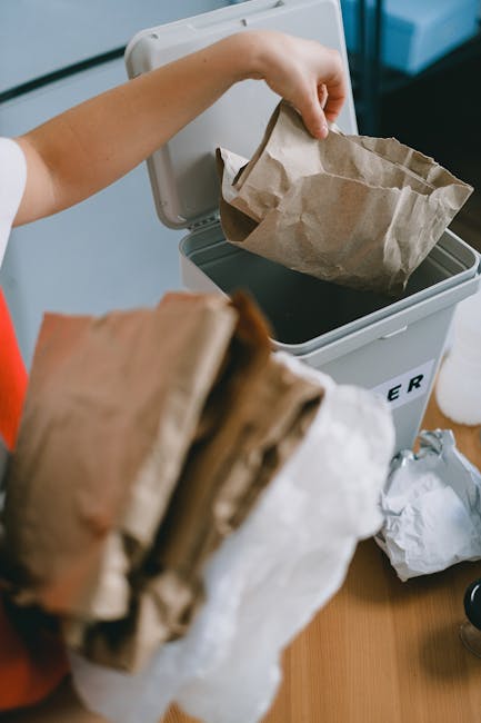 A person is disposing of a crumpled brown paper bag into a white recycling bin, which is placed on a wooden surface. The individual, wearing a white shirt, is seen from the arm up, with their hand hol