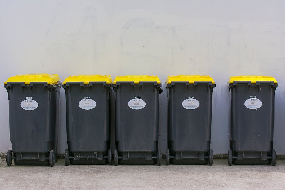 A person is disposing of a crumpled brown paper bag into a white recycling bin, which is placed on a wooden surface. The individual, wearing a white shirt, is seen from the arm up, with their hand hol